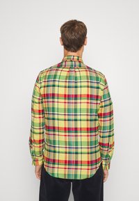 Zadní pohled na flanelovou košili s dlouhým rukávem v žluté, červené, zelené a modré barvě. Měkká tkanina, standardní límeček a knoflíkové manžety. Zaoblený lem.