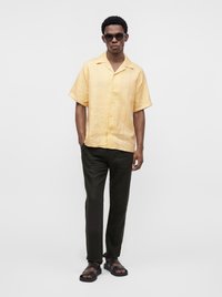 Homme portant des lunettes de soleil, une chemise jaune à manches courtes boutonnée, un pantalon foncé et des sandales noires, debout devant un fond clair uni.
