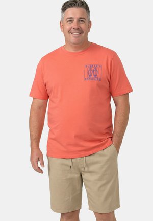 Lächelnder Mann mit korallenfarbenem T-Shirt mit "Surfer's Paradise"-Logo und beigen Shorts, der mit einer Hand in der Tasche vor einem einfarbigen Hintergrund steht.