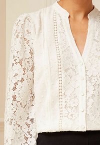 Bílá krajková blůza s nabranými rukávy, mandarínkovým límcem, knoflíkovým zapínáním vpředu a květinovým vzorem s texturovanými akcenty.