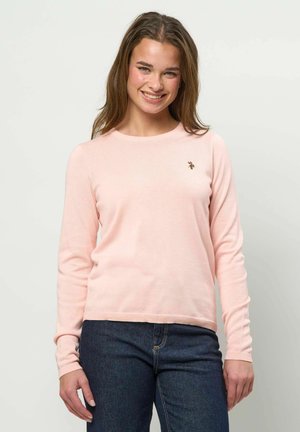 Joven sonriente, vestida con un suéter de manga larga rosa claro y jeans azul oscuro, posando frente a un fondo liso y claro.