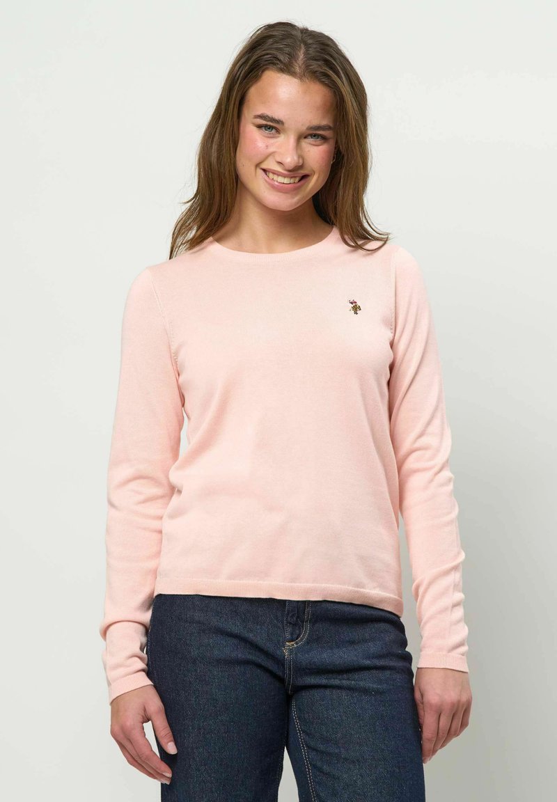 Jeune femme souriante, portant un pull léger à manches longues rose pâle et un jean bleu foncé, posant devant un fond clair uni.