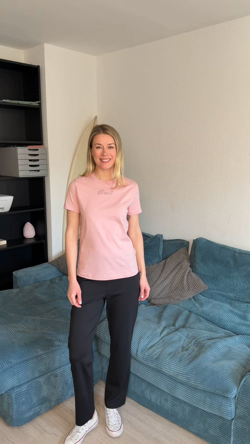 Млада жена с руса коса, облечена в розова тениска и черни панталони, стои до син ъглов диван в модерен хол.