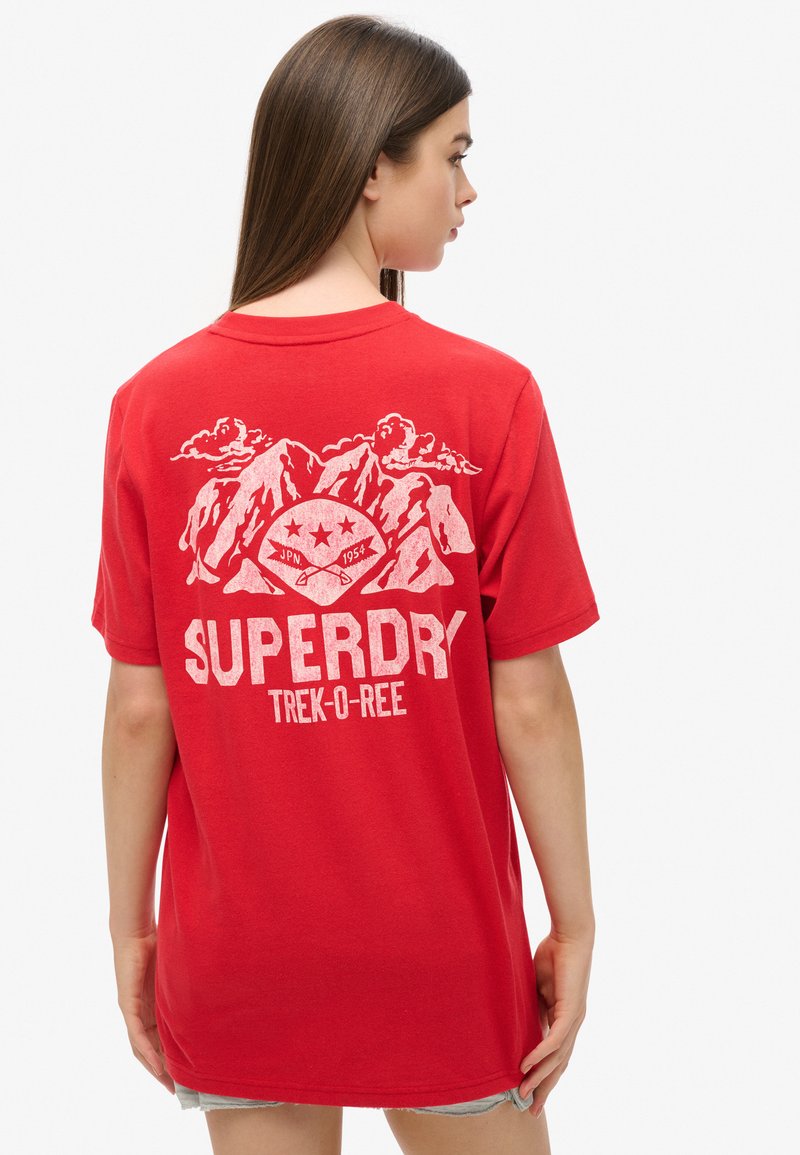 Femme aux longs cheveux bruns portant un T-shirt rouge Superdry avec un motif blanc de montagne et de pagaie au dos.