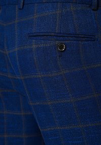 Niebieskie spodnie z tkaniny o fakturowym splocie, z delikatnym brązowym wzorem kraty. Zawierają tylną kieszeń z pojedynczym czarnym guzikiem.