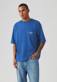 Μπλε βαμβακερό T-shirt με κοντά μανίκια, διαθέτει τσέπη στο στήθος και λευκή κεντημένη επιγραφή. Φορεμένο με ανοιχτό μπλε τζιν.