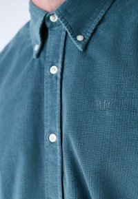 Cămașă cu un model texturat, de culoare verde închis, cu guler, butoane albe și un logo brodat subtil pe piept.