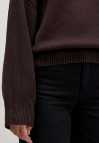 Brauner strukturierter Strickpullover mit langen Ärmeln und geripptem Saum, kombiniert mit schwarzen Hosen. Der Stoff wirkt weich und warm.