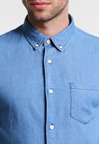 Camicia in denim azzurro chiaro con colletto a bottoni, chiusura frontale con bottoni, tasca sul petto e una sottile trama.