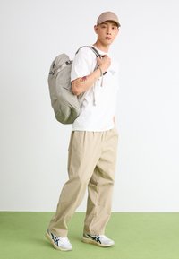 Light gray backpack with multiple pockets, worn on a man's back. He wears a white t-shirt, beige pants, and gray sneakers with navy accents.