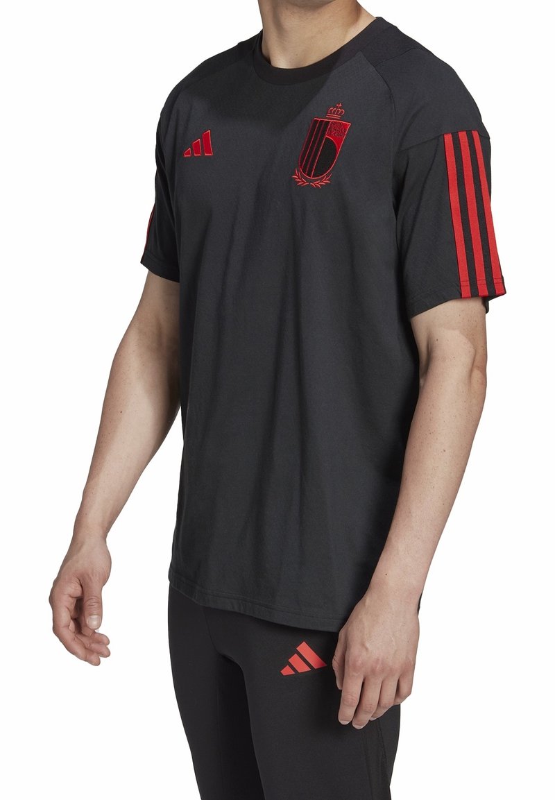 T-shirt de sport noir avec des bandes rouges Adidas sur les manches, arborant un logo bouclier rouge sur la poitrine. Tissu doux, coupe décontractée.