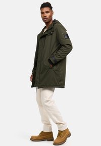 Green parka with a front zipper, hood, and multiple pockets. Paired with cream pants and brown lace-up boots. Smooth fabric finish.