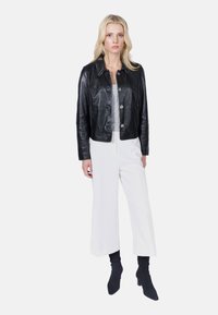 Černá kožená zkrácená bunda s lesklým povrchem, vybavená knoflíkovým zapínáním a límečkem, v kombinaci s bílými širokými kalhotami.