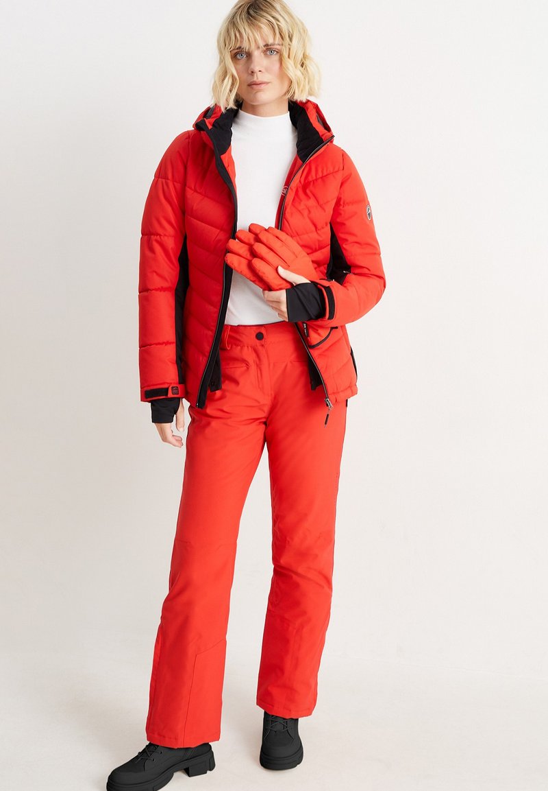 Rote isolierte Daunenjacke mit schwarzen Akzenten, passende rote Skihosen und orangefarbene Handschuhe; schwarze Schuhe vervollständigen das Outfit.