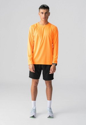 Ung mand stående, iført orange langærmet skjorte, sorte shorts, hvide sokker, lysegrå sneakers, solbriller på hovedet og smartwatch på håndleddet.