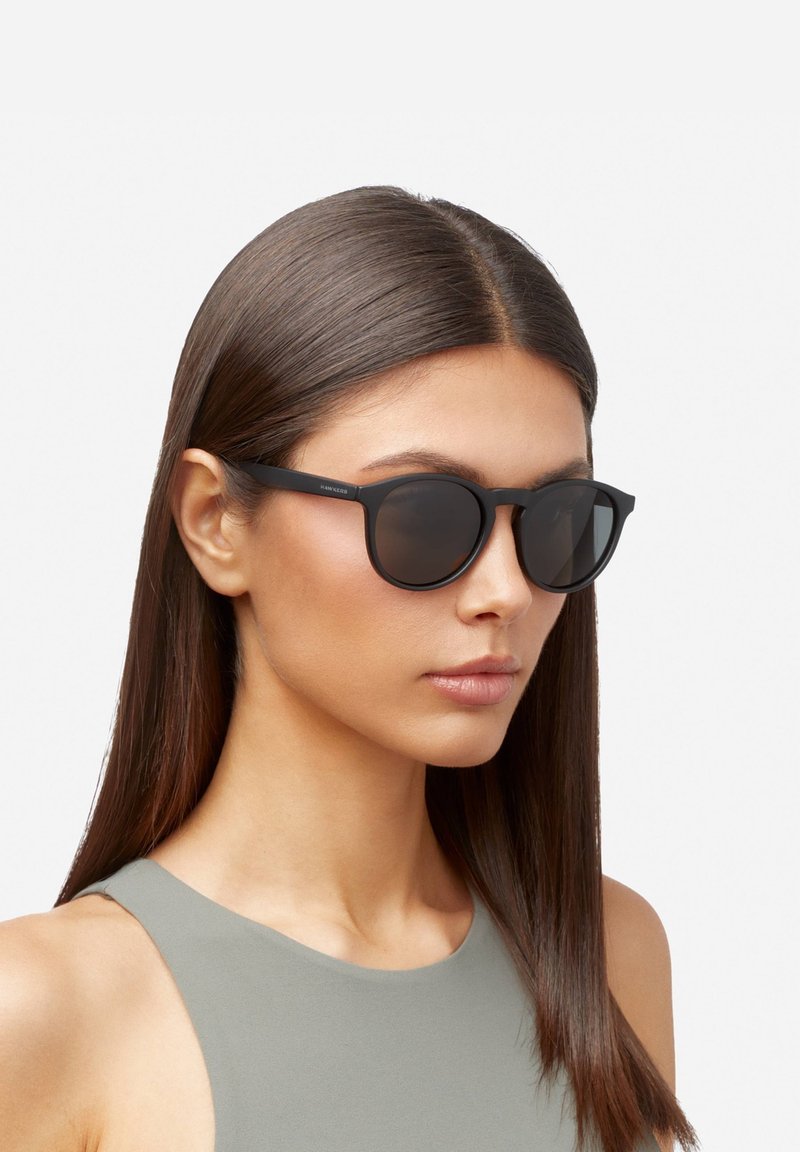 Occhiali da sole neri con montatura rotonda, texture opaca e lenti scure. Indossati da una modella con capelli castano chiaro, lunghi e lisci.