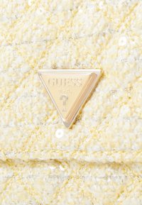 Bolso de mano de tela amarilla acolchada que presenta hilos plateados, acentos de lentejuelas y un logotipo triangular de Guess en dorado.