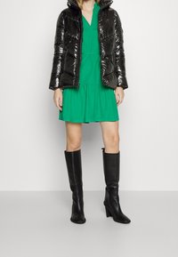 Chaqueta de plumas negra, vestido verde, botas de tacón negras hasta la rodilla. Textura suave, detalle de cremallera en la chaqueta, silueta en A en el vestido.