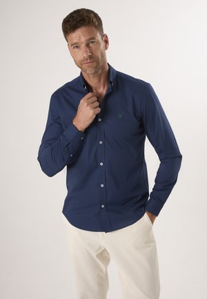 Marineblaue Langarmbluse mit Button-Down-Kragen, weißen Knöpfen und einem grünen Logo-Akzent auf der Brust, kombiniert mit cremefarbenen Hosen.