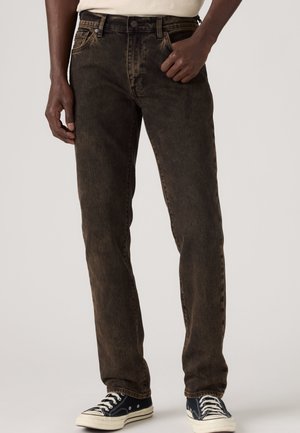 Homme portant un jean droit marron foncé et des baskets basses noires avec des lacets blancs, debout devant un fond uni.