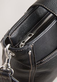 Sac en cuir noir avec fermeture éclair argentée partiellement ouverte, montrant des coutures détaillées et une boucle métallique sur la bandoulière réglable.