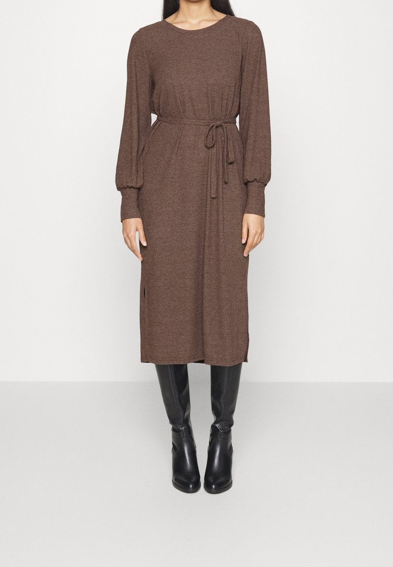 Femme portant une robe midi marron à manches longues avec une ceinture, associée à des bottes en cuir noires montant jusqu'aux genoux, debout devant un fond uni.