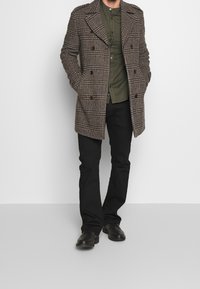 Abrigo de lana en diseño de espiga marrón y negro con doble botonadura, grandes solapas y dos bolsillos laterales, llevado sobre una camisa verde oliva y pantalones negros.