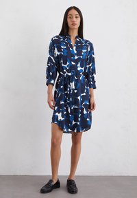 Abito camicia con uno sfondo blu navy caratterizzato da motivi astratti bianchi e blu. Maniche lunghe, chiusura a bottoni e vita con cintura.