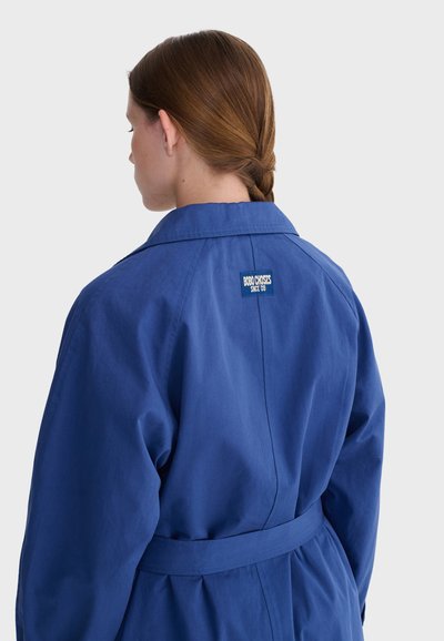 Femme aux cheveux bruns tressés portant un manteau bleu roi ceinturé avec une étiquette "Bobo Choses Since '09" sur le haut du dos, vue de dos.