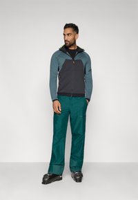 Pánsky outdoorový outfit pozostávajúci z tej modrozelenej a tmavosivej bundy so zipsom a modrozelenej nohavice. Odolný materiál s funkčným dizajnom a akcentmi.