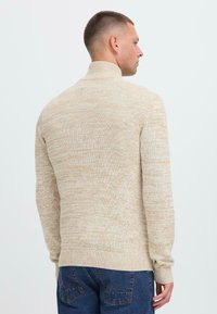 Mann som vender bort med en beige melert genser med høy hals og blåjeans mot en ensfarget lys bakgrunn.