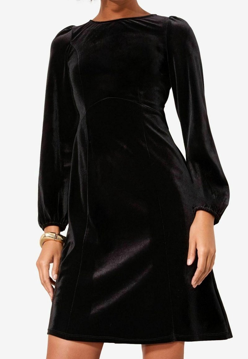Robe en velours noir longueur genou avec manches longues bouffantes et taille cintrée, portée par une personne debout avec un bracelet en or au poignet gauche.