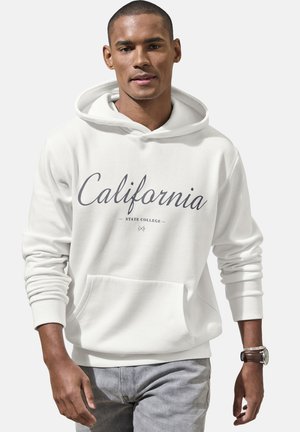 Junger Mann, der einen weißen Hoodie mit der Aufschrift "California State College", hellgraue Jeans und eine Armbanduhr mit braunem Lederarmband trägt.