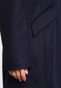 Marinblå ullrock med en stilren design, synliga fickdetaljer och en mjuk textur, med en hand som vilar på ärmen för skala.