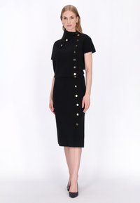 Frau trägt ein schwarzes Kurzarmkleid mit diagonalen goldenen Knöpfen von der Schulter bis zum Saum, kombiniert mit schwarzen spitzen High Heels.