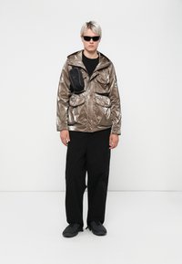 Jachetă zipuită metalică argintie cu multiple buzunare, combinată cu pantaloni cargo negri largi și încălțăminte neagră. Accesoriile includ ochelari de soare.