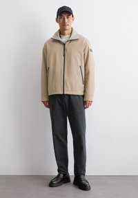 Beige Zip-Jacke mit hohem Kragen und zwei vorderen Taschen, kombiniert mit dunkelgrauen Hosen und schwarzen Schuhen. Glatter Stoff mit minimalem Design.