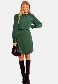 Zelena obleka z dolgimi rokavi in visokim ovratnikom, zategnjeno v pasu in z rahlim sijajem, kombinirana s črno torbico in črnimi čevlji do kolen.