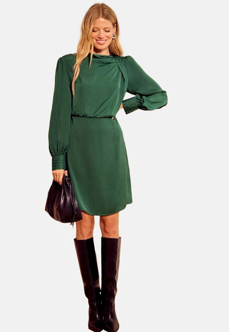 Zelena obleka z dolgimi rokavi in visokim ovratnikom, zategnjeno v pasu in z rahlim sijajem, kombinirana s črno torbico in črnimi čevlji do kolen.