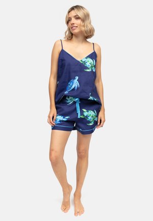 Vrouw draagt een marineblauwe camisole en korte broek met blauwgroene zeeschildpaddenprint, staat blootsvoets op een lichte achtergrond.