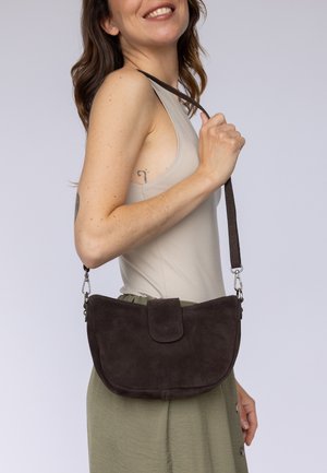 Frau trägt beige Oberteil und grünen Rock, dazu eine dunkelbraune Wildleder-Schultertasche mit Klappe.