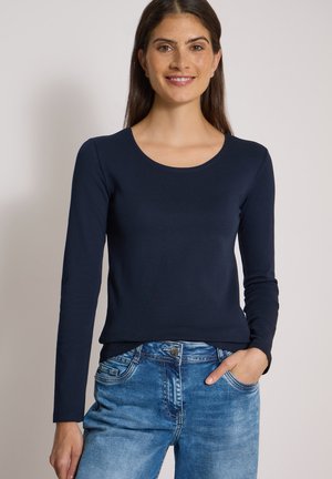 Femme aux longs cheveux bruns portant une chemise bleu marine à manches longues et un jean bleu, souriant avec une main dans la poche, sur un fond uni.