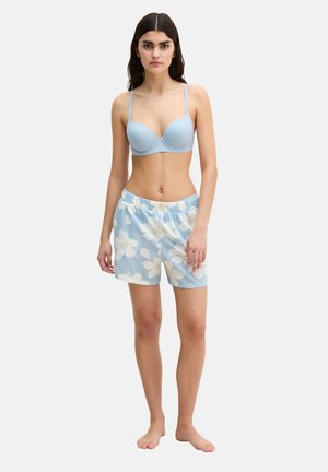 Kobieta stojąca boso, ubrana w jasnoniebieski stanik i pasujące kwieciste szorty na gładkim tle.