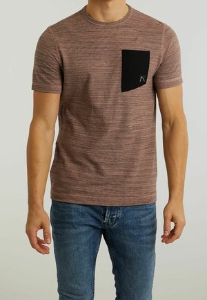 Mężczyzna noszący brązowy, krótkim rękawem teksturowany t-shirt z czarną prostokątną kieszenią na piersi, w połączeniu z niebieskimi dżinsami.