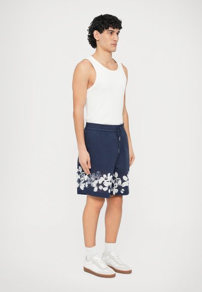 Shorts bleu marine avec imprimé floral blanc à l'ourlet, taille élastique, poches latérales, assortis à un débardeur blanc et des baskets blanches.
