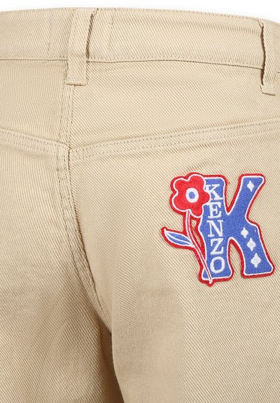 Pantalon en toile beige présentant une surface texturée, avec un patch brodé coloré affichant le nom "KENZO" et des accents floraux.
