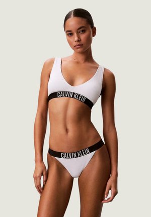 Donna che indossa un bralette in tessuto costina bianco Calvin Klein e biancheria intima coordinata con bande elastiche nere con il nome del marchio.
