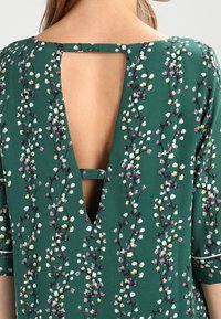 Vestido verde con un patrón floral que presenta acentos en púrpura y blanco, diseño de espalda descubierta con una cinta horizontal y mangas cortas.