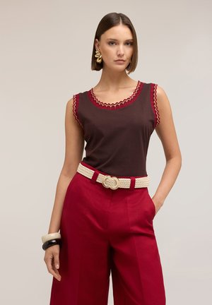 Kobieta ubrana w brązową bluzkę bez rękawów z czerwonym wykończeniem, czerwone, wysokie spodnie z plecionym kremowym paskiem, złote kolczyki oraz czarno-kremowe bransoletki.