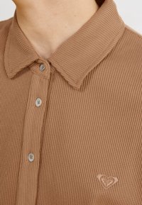 Polo marrón de canalé con un acabado texturizado, que presenta un collar, cierre de botones y un pequeño logo en forma de corazón bordado en el pecho.